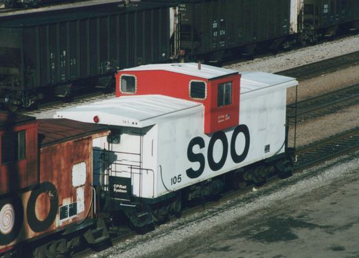 A white caboose with large black 'SOO' lettering and a red cupola, numbered 105, sitting on railroad tracks in a yard with dark green freight cars behind it.