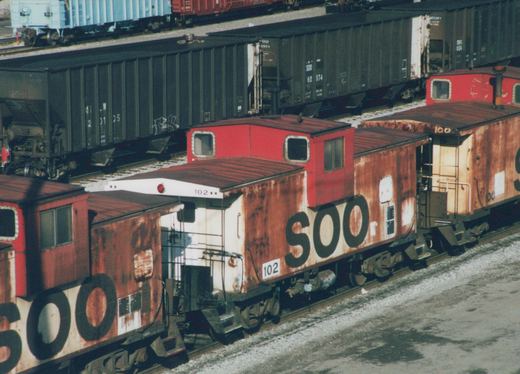 Two rusted SOO Line cabooses are parked on parallel tracks in a snowy rail yard, with a boxcar behind them.