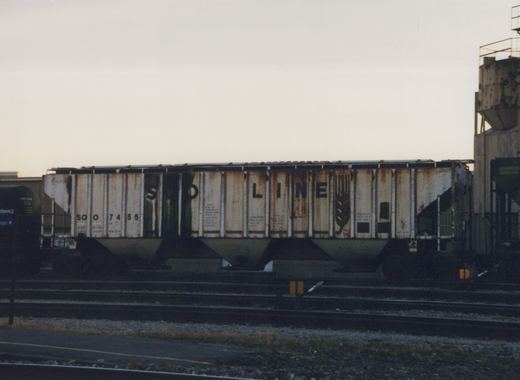 A side view of a Soo Line freight car with the number 74553, parked on multiple tracks in a rail yard at dusk with a platform sign visible on the left.
