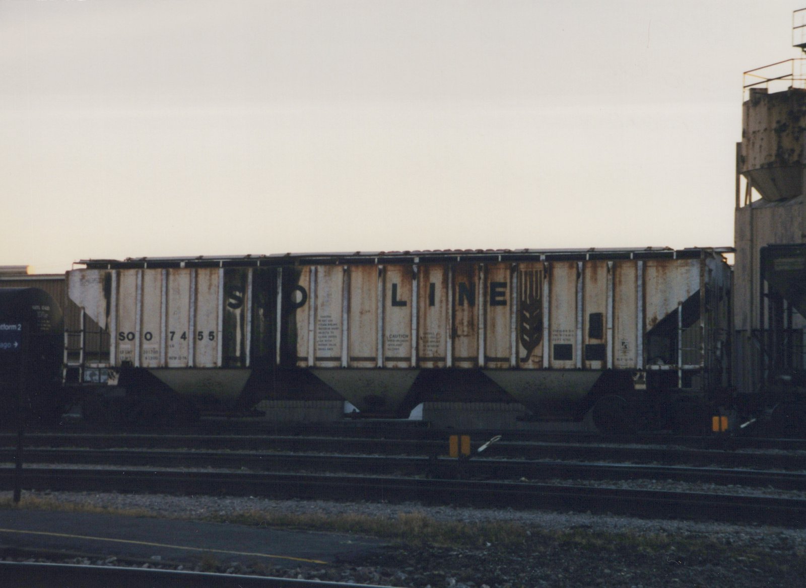 A side view of a Soo Line freight car with the number 74553, parked on multiple tracks in a rail yard at dusk with a platform sign visible on the left.
