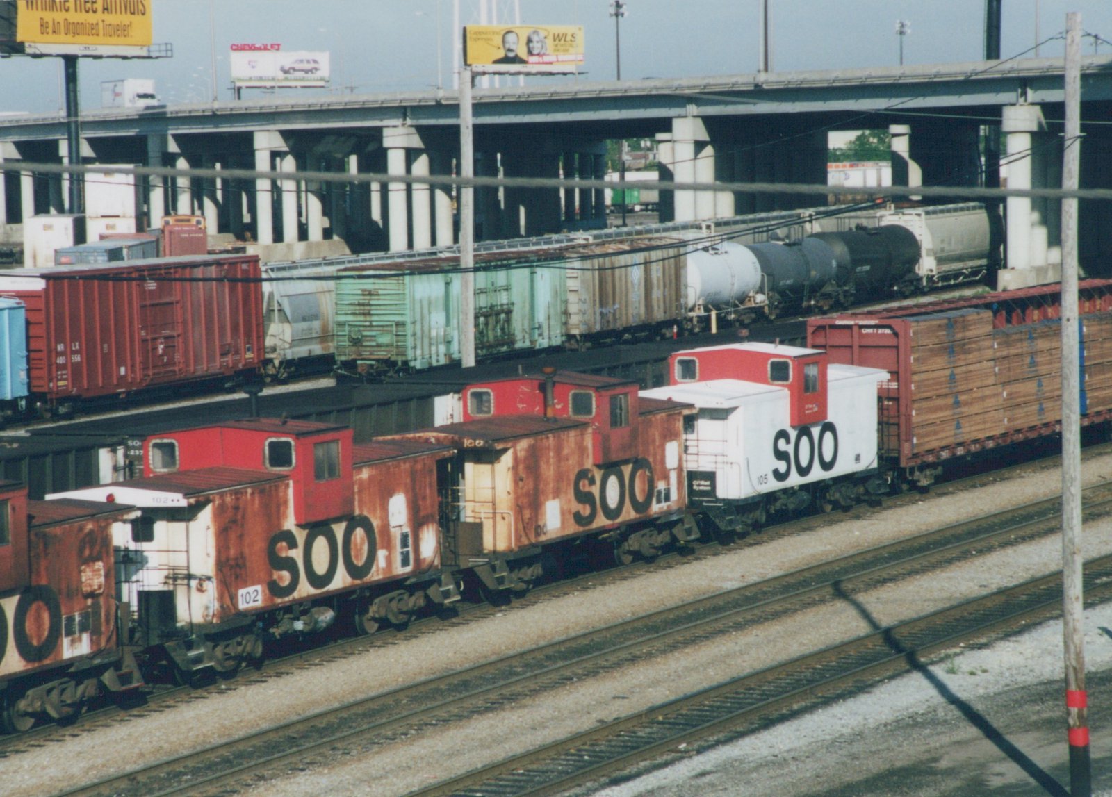A row of rusted and white Soo Line cabooses, numbered 102 and 105, sit on tracks in a busy rail yard under a concrete overpass, with multiple freight cars visible in the background.