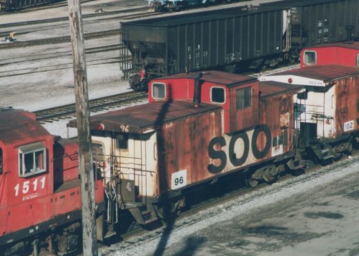 A red and white SOO Line caboose numbered 96 is parked on a railway track in a yard, with a red CP Rail locomotive numbered 1511 beside it.