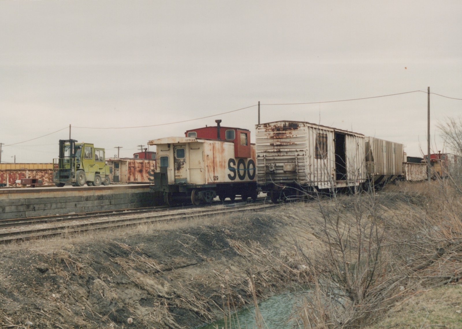 A side view of a weathered Soo Line caboose with number 129 parked on a track in a yard with old freight cars and a forklift nearby, during a winter day.