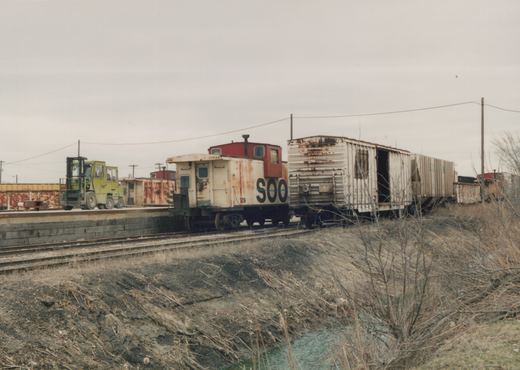 A side view of a weathered Soo Line caboose with number 129 parked on a track in a yard with old freight cars and a forklift nearby, during a winter day.