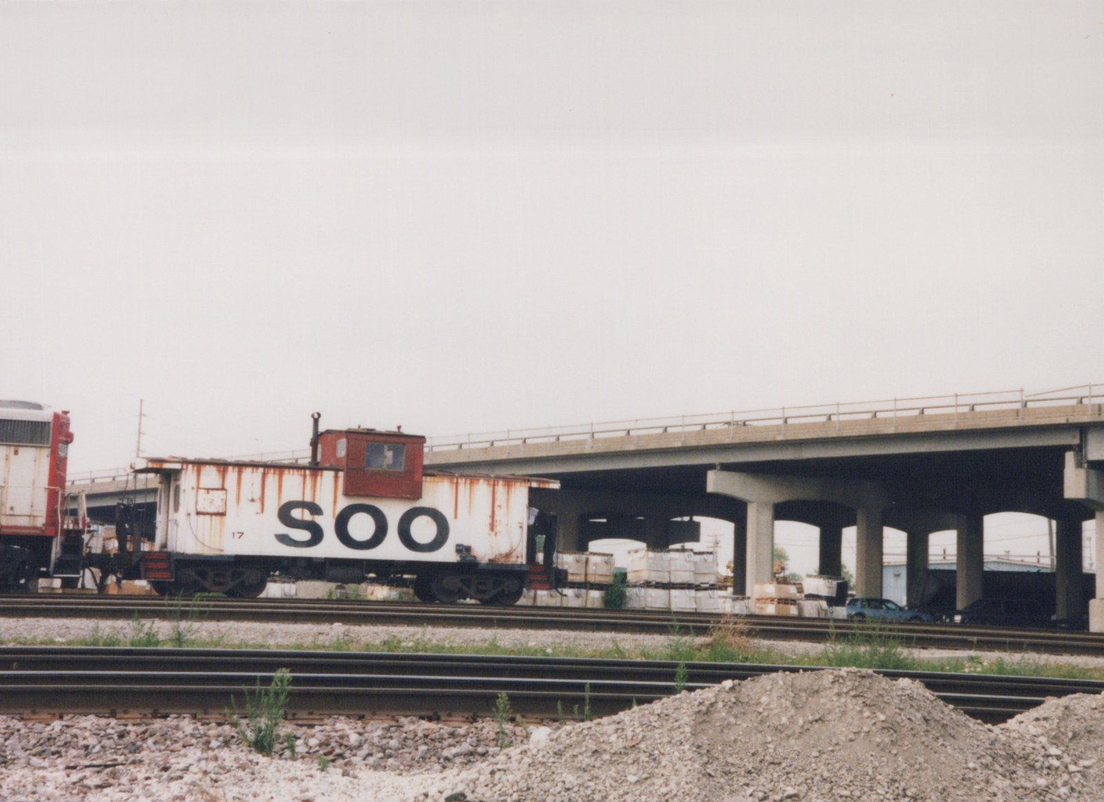 A white and red SOO Line caboose numbered 17 sits on railroad tracks under a large concrete bridge on a cloudy day, with piles of gravel in the foreground.