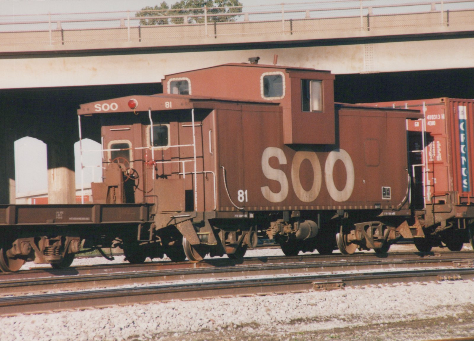 A brown SOO Line caboose numbered 81 sits on a railroad track under a concrete bridge, with a freight car behind it.