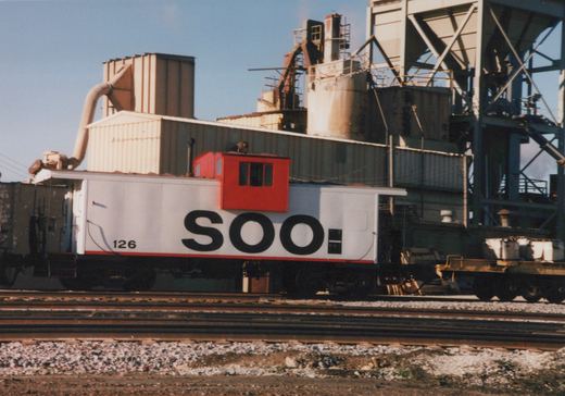 A white and red Soo Line caboose, number 126, is parked on railroad tracks next to an industrial building under a clear sky.