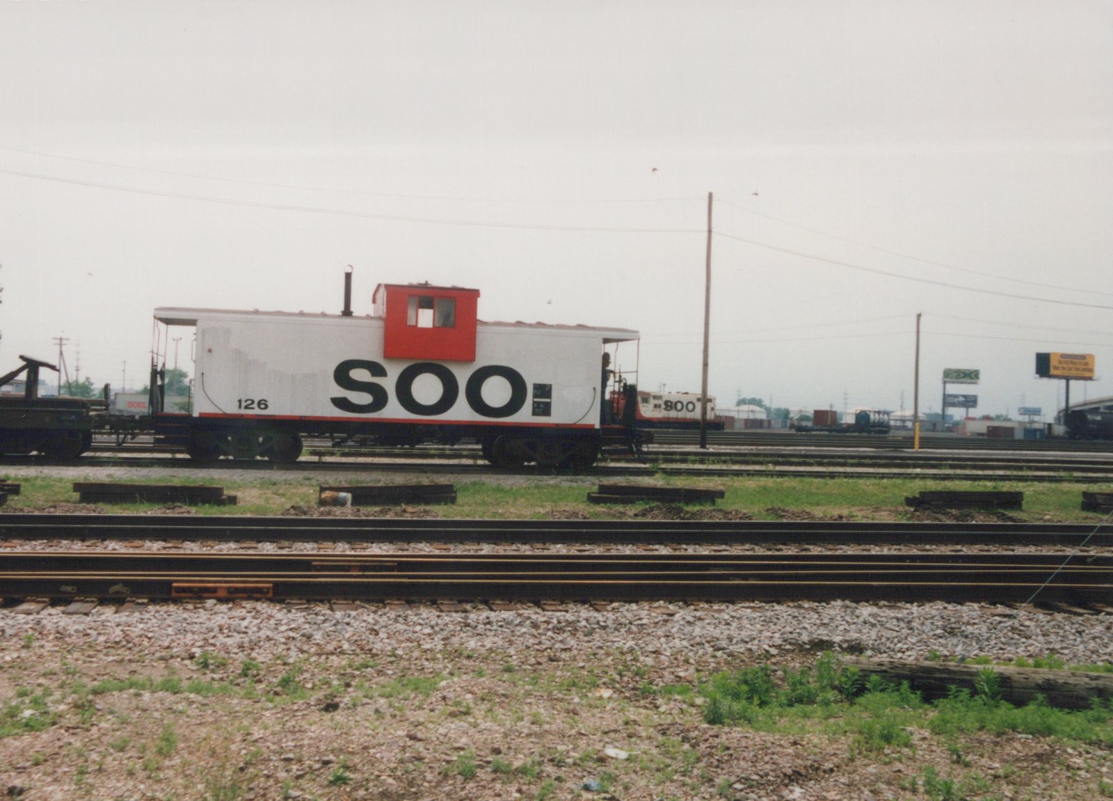 A white SOO Line caboose with the number 126 is parked on tracks in a rail yard, with a person on its rear platform and a SOO Line locomotive #800 visible in the background.