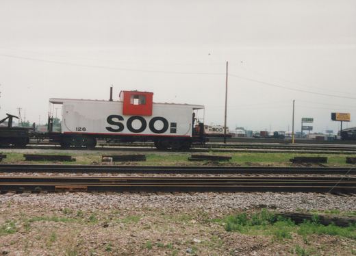 A white SOO Line caboose with the number 126 is parked on tracks in a rail yard, with a person on its rear platform and a SOO Line locomotive #800 visible in the background.
