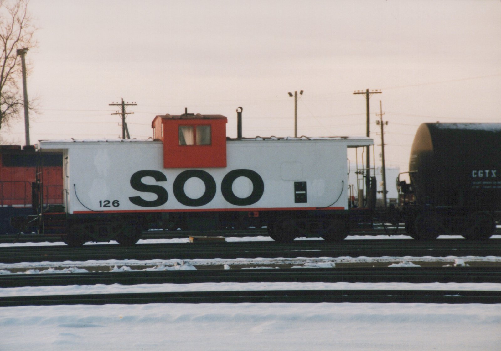 White SOO Line caboose with red cupola, number 126, parked on snowy tracks beside a black tank car in a railroad yard under an overcast sky.