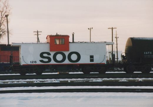 White SOO Line caboose with red cupola, number 126, parked on snowy tracks beside a black tank car in a railroad yard under an overcast sky.