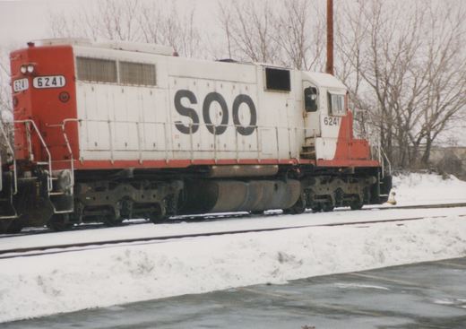 A white and red Soo Line diesel locomotive numbered 6241 sits on tracks beside a snow-covered embankment with bare trees in the background.