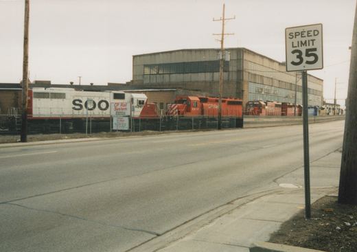 A street view of a large industrial building with several diesel locomotives in SOO Line and CP Rail livery parked in a rail yard behind a fence, with a 'SPEED LIMIT 35' sign in the foreground.