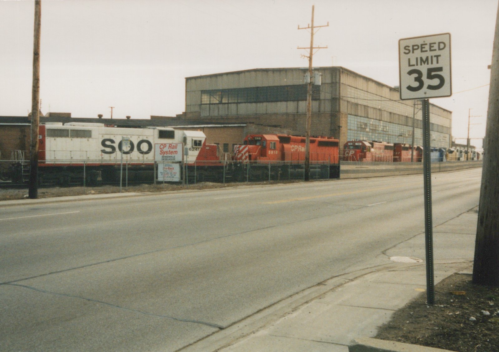 A street view of a large industrial building with several diesel locomotives in SOO Line and CP Rail livery parked in a rail yard behind a fence, with a 'SPEED LIMIT 35' sign in the foreground.