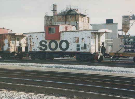 A side view of a white and rusty Soo Line caboose numbered 63, parked on railroad tracks in an industrial yard.