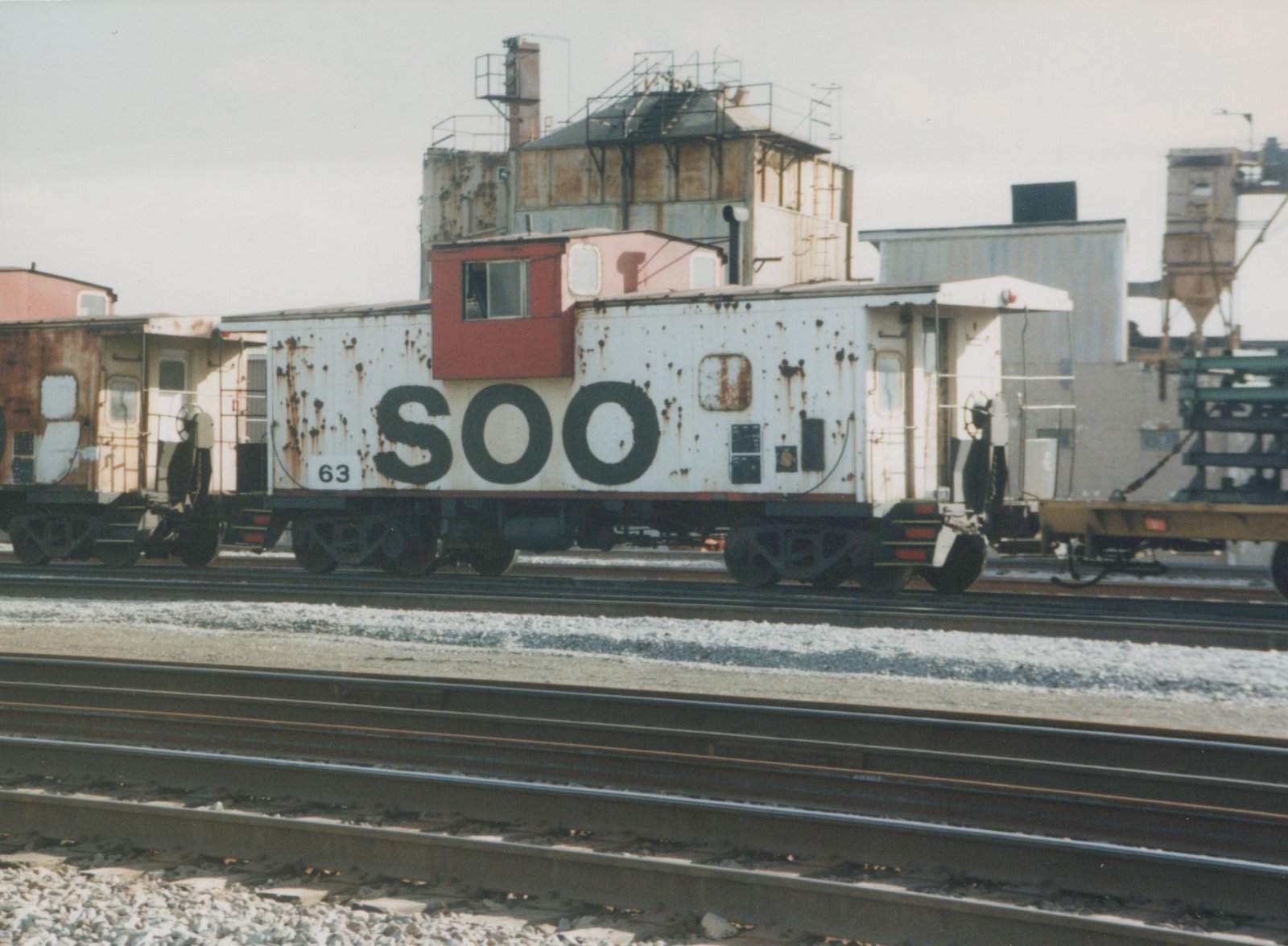 A side view of a white and rusty Soo Line caboose numbered 63, parked on railroad tracks in an industrial yard.
