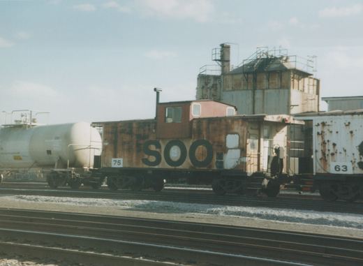 A side view of a rusty brown SOO Line caboose, number 75, in a rail yard with multiple tracks, a white tank car, and a rusted white boxcar nearby.