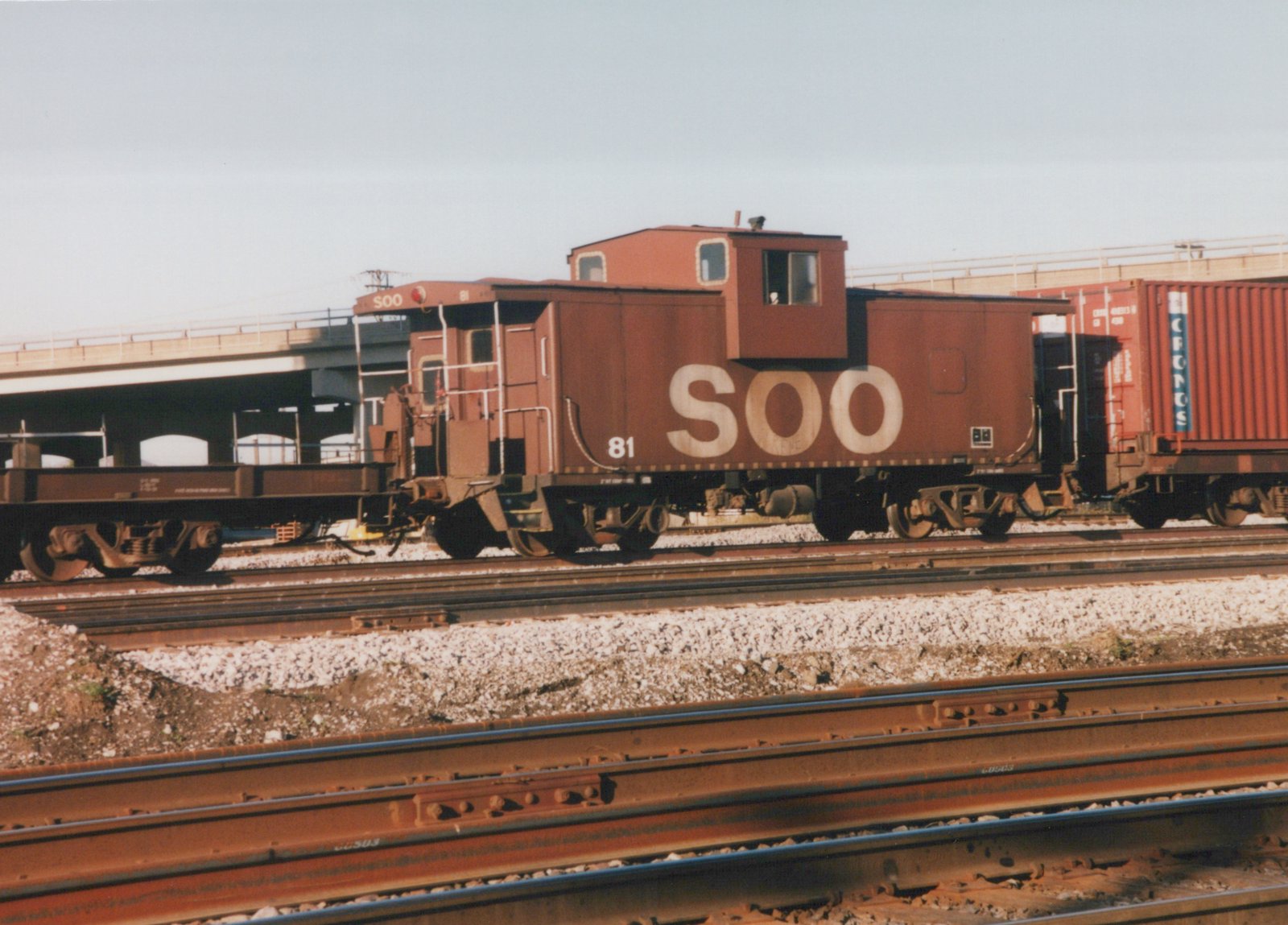 A maroon Soo Line caboose numbered 81 is visible on a set of railroad tracks, with a shipping container car behind it and a bridge in the background.