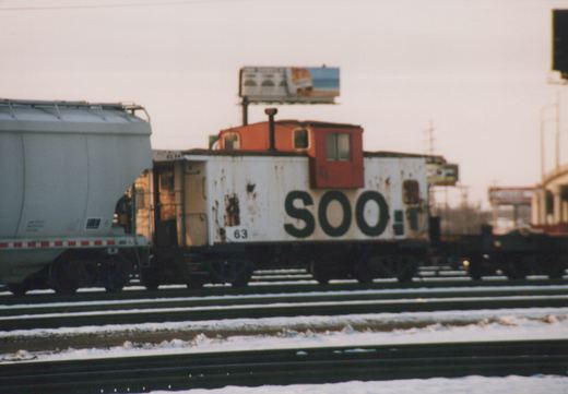 A side view of a white Soo Line caboose with a red cupola, numbered 63, parked on a snowy railroad track next to a covered hopper car.