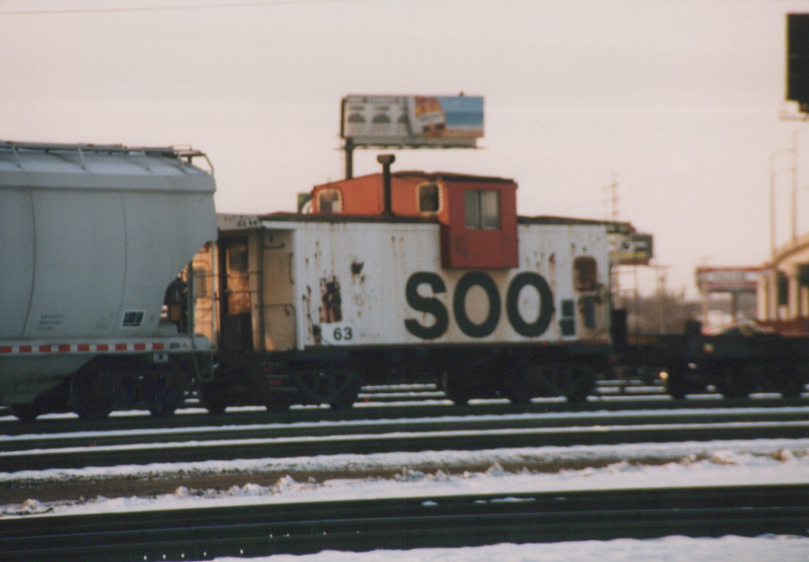 A side view of a white Soo Line caboose with a red cupola, numbered 63, parked on a snowy railroad track next to a covered hopper car.