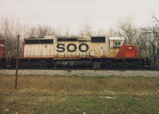 Side view of a Soo Line diesel locomotive, number 6608, painted in white and red, on a railroad track with bare trees in the background on an overcast day.