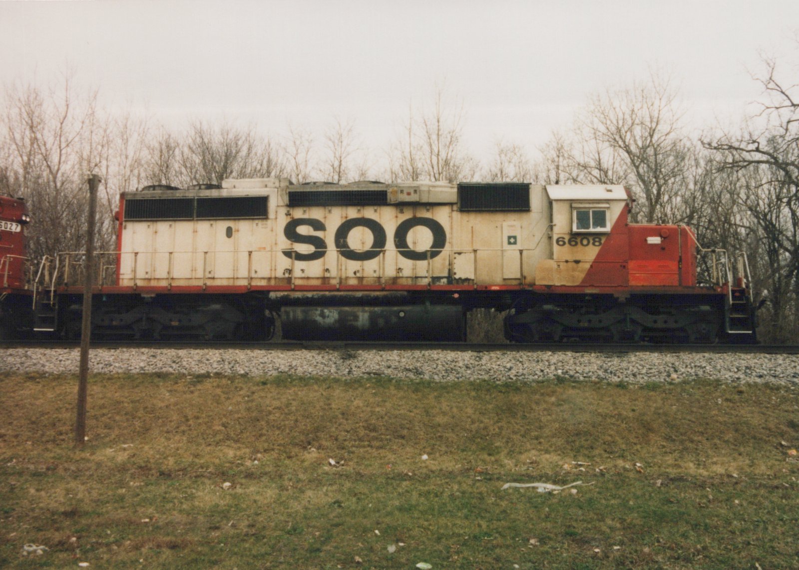 Side view of a Soo Line diesel locomotive, number 6608, painted in white and red, on a railroad track with bare trees in the background on an overcast day.