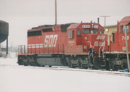 A red SOO Line diesel locomotive numbered 6604 is parked on snowy tracks, with a person in an orange hat standing on its steps. Another red SOO train is next to it, and a water tower is visible in the background.