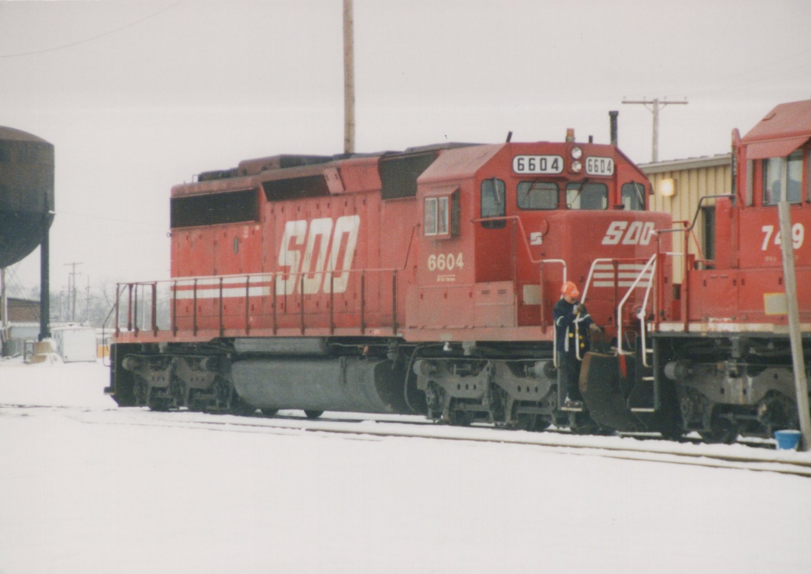A red SOO Line diesel locomotive numbered 6604 is parked on snowy tracks, with a person in an orange hat standing on its steps. Another red SOO train is next to it, and a water tower is visible in the background.