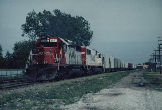 A red and white SOO Line diesel locomotive numbered 6602 pulls a long train of freight cars on a track beside a gravel road, with trees and utility poles in the background under an overcast sky.