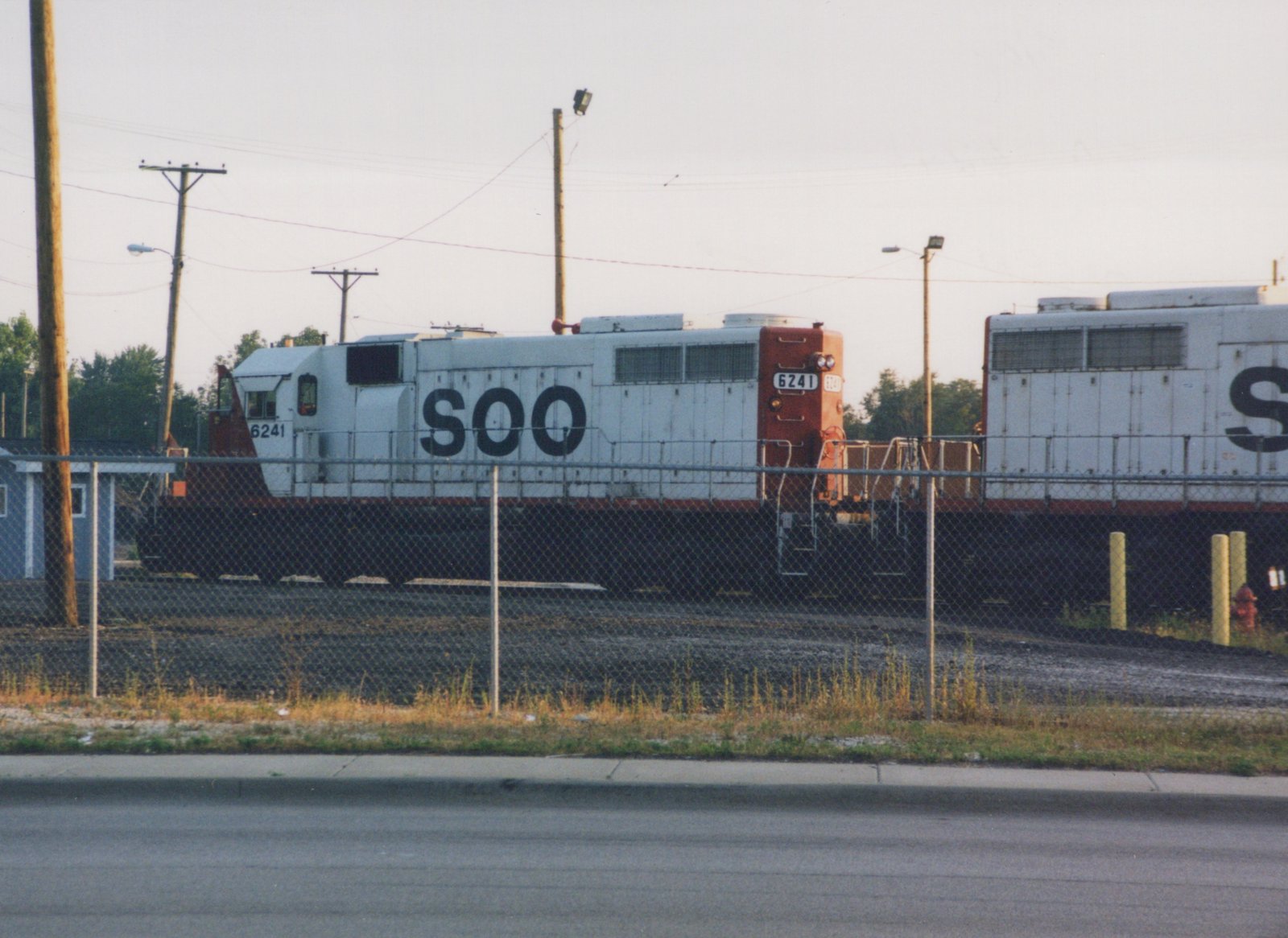 A side view of two white Soo Line diesel locomotives, including number 6241, parked on tracks behind a chain-link fence on an overcast day.