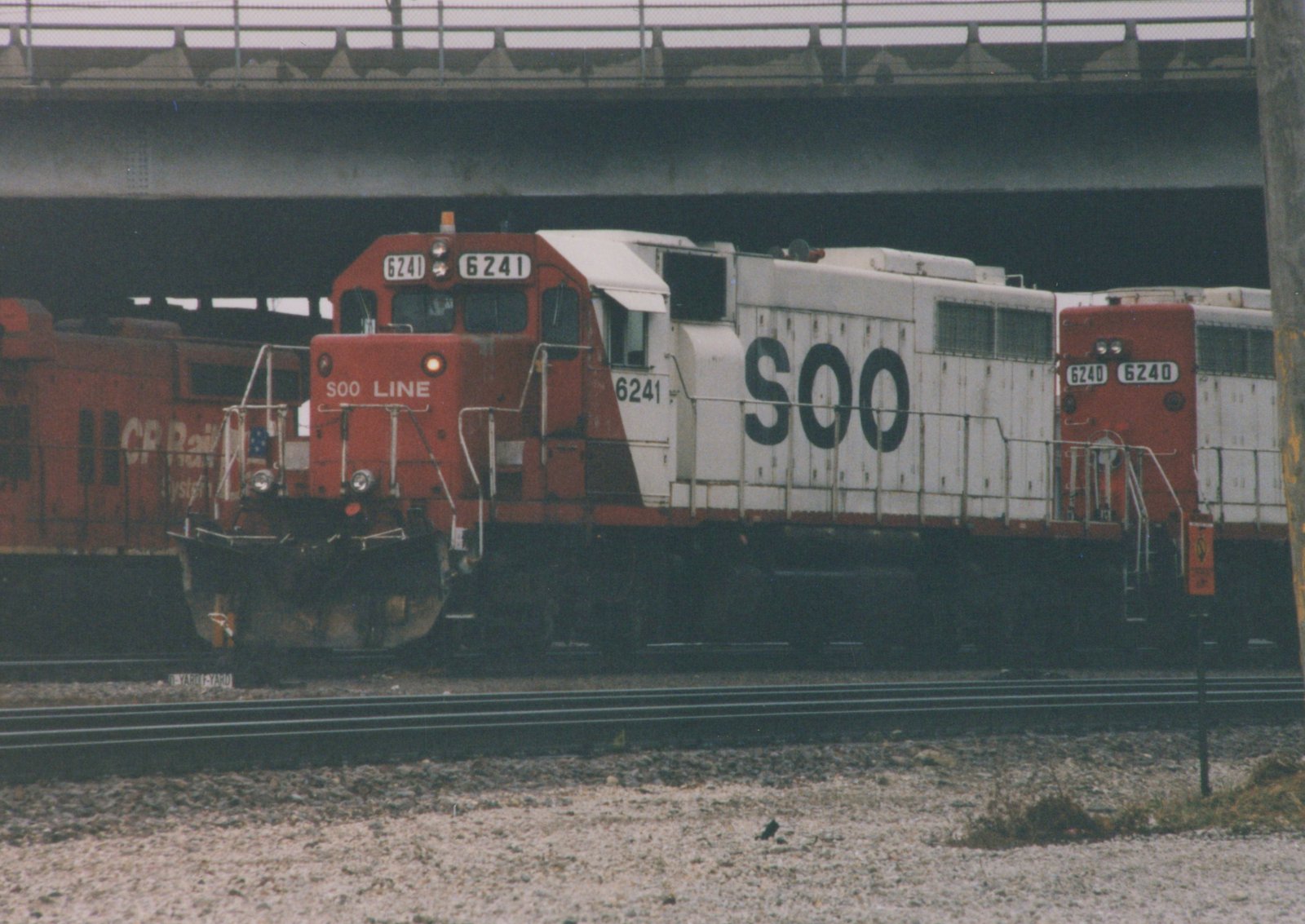 Two red and white SOO Line diesel locomotives, numbered 6241 and 6240, stand on tracks beneath an overpass in a rail yard, with another red locomotive behind them.