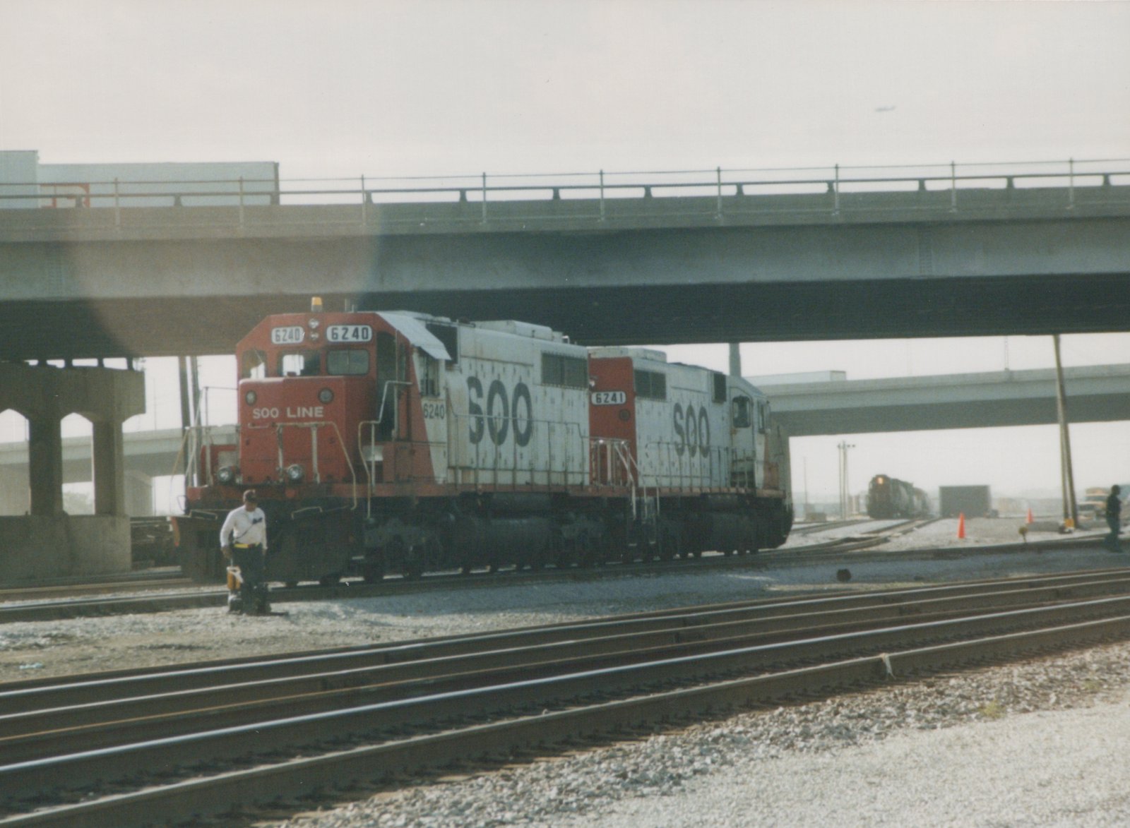 Two white and red SOO Line diesel locomotives numbered 6240 and 6241 are parked on railroad tracks under a large concrete overpass, with two people visible nearby on a hazy day.