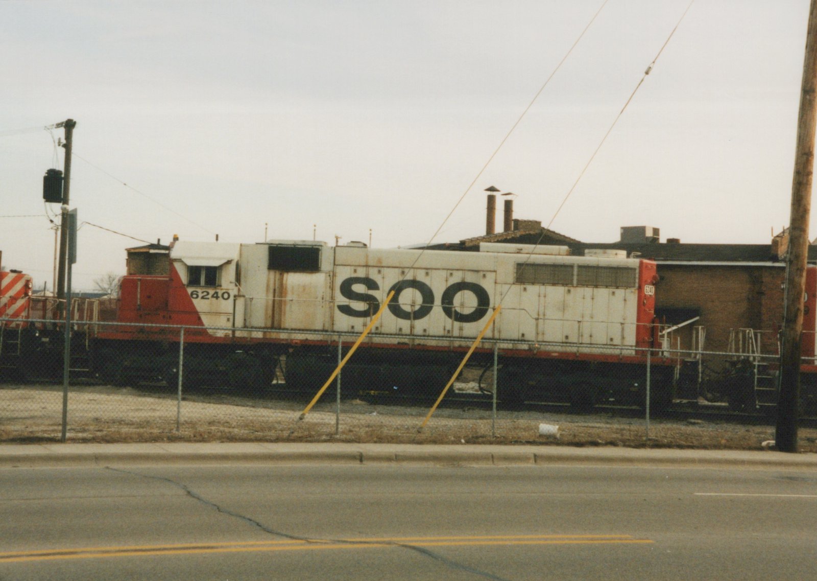 Side view of Soo Line diesel locomotive 6240, white with red ends and black lettering, parked on tracks behind a fence on a cloudy day.