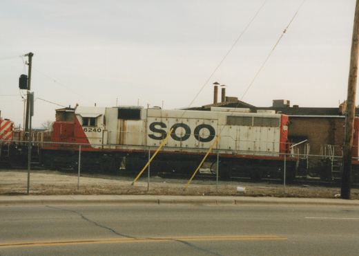 Side view of Soo Line diesel locomotive 6240, white with red ends and black lettering, parked on tracks behind a fence on a cloudy day.