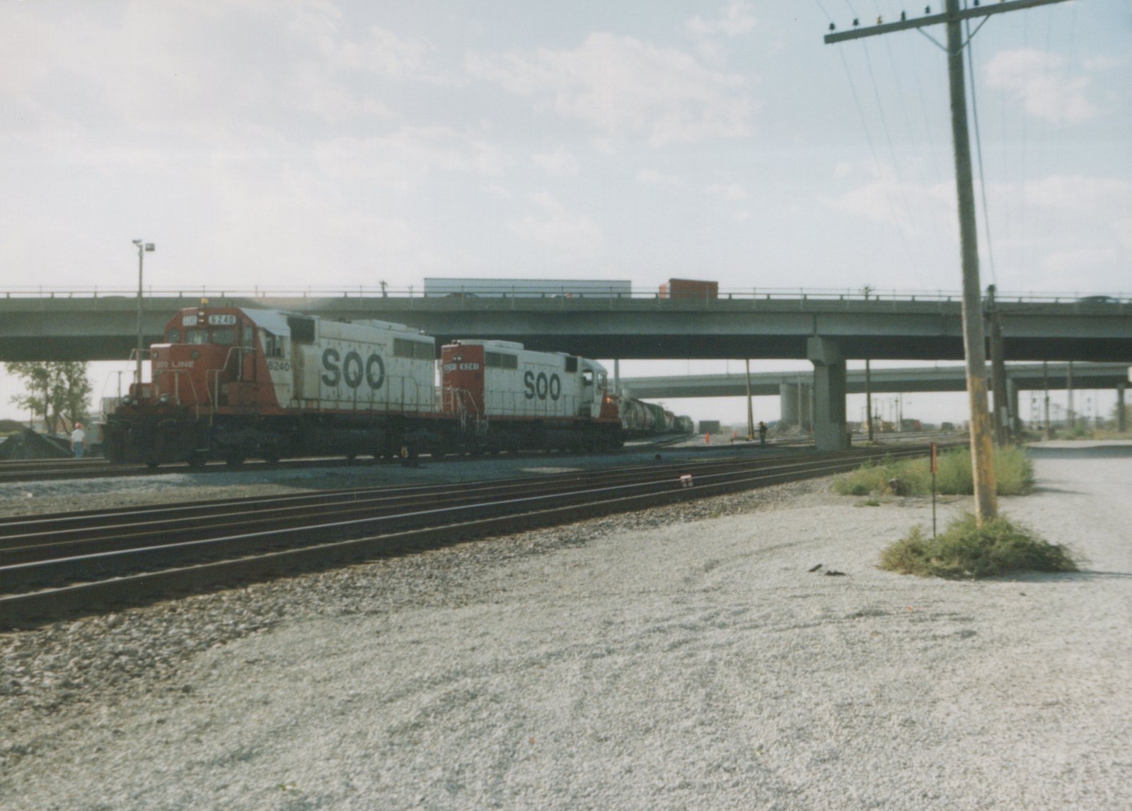 Two SOO Line diesel locomotives, numbered 6240 and 6241, are coupled together, pulling a train of freight cars under a highway overpass on a sunny day.