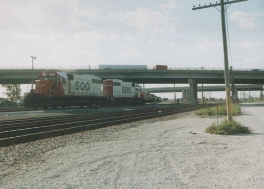 Two SOO Line diesel locomotives, numbered 6240 and 6241, are coupled together, pulling a train of freight cars under a highway overpass on a sunny day.