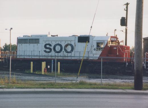 A white and red SOO Line diesel locomotive numbered 6240, viewed from a three-quarter front angle behind a chain-link fence in a yard on an overcast day.