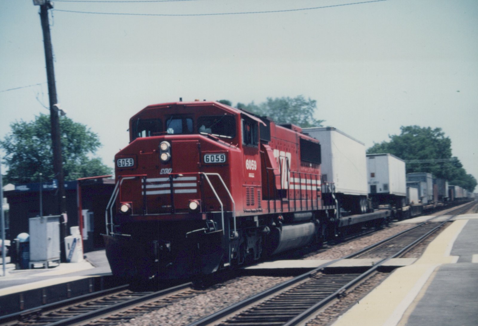 A red SOO Line locomotive numbered 6059 pulls a long train of white trailers on flatcars along a railroad track beside a concrete platform.