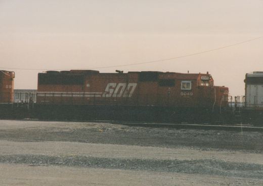 A side view of an orange SOO Line diesel locomotive numbered 6049, parked on a track in a gravel yard under an overcast sky.