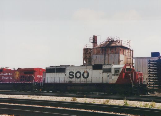 Two diesel locomotives, one white with red cab marked SOO 6036 and another red marked Canadian Pacific 8534, are coupled together on tracks in a yard with industrial buildings in the background.
