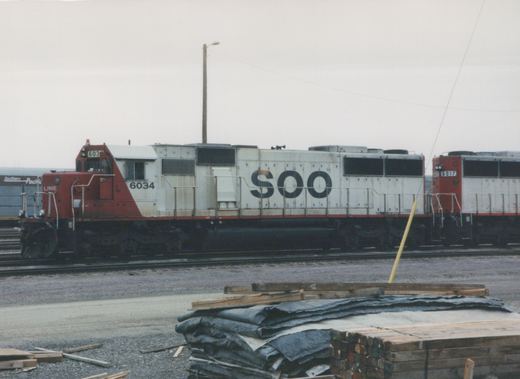 A side view of a white and red SOO Line diesel locomotive, number 6034, parked in a gravel rail yard with another locomotive and a freight car behind it.