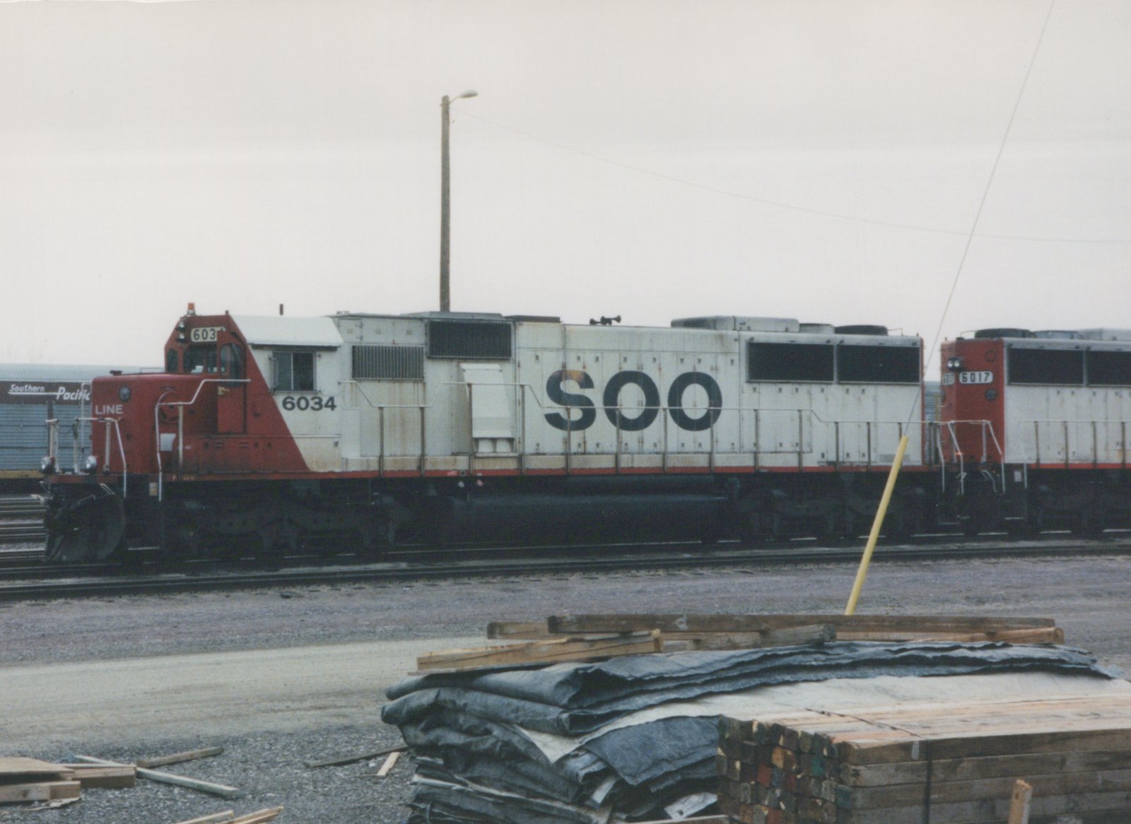 A side view of a white and red SOO Line diesel locomotive, number 6034, parked in a gravel rail yard with another locomotive and a freight car behind it.