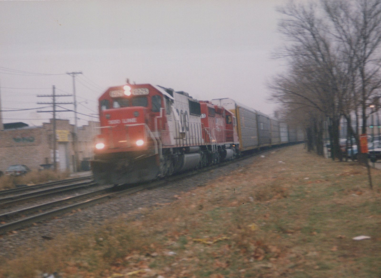 A red and white SOO Line diesel locomotive, number 8025, travels down a railroad track with a line of freight cars, viewed from a three-quarter front angle on an overcast autumn day.