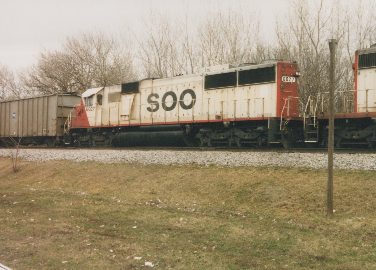A white and red Soo Line locomotive numbered 6027 is on train tracks beside a grassy area with bare trees in the background.