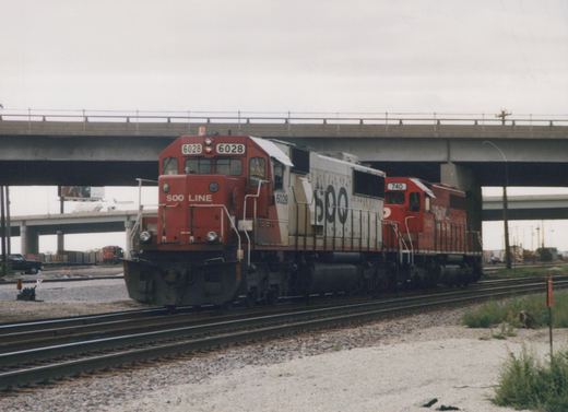 A red and white SOO Line locomotive numbered 6028 leads a red locomotive numbered 740 on railroad tracks beneath a concrete overpass on a cloudy day.