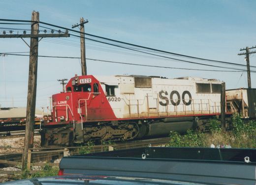A red and white Soo Line diesel locomotive numbered 6020 is shown on a railroad track under a clear blue sky, with a car roof in the foreground and a freight car in the background.
