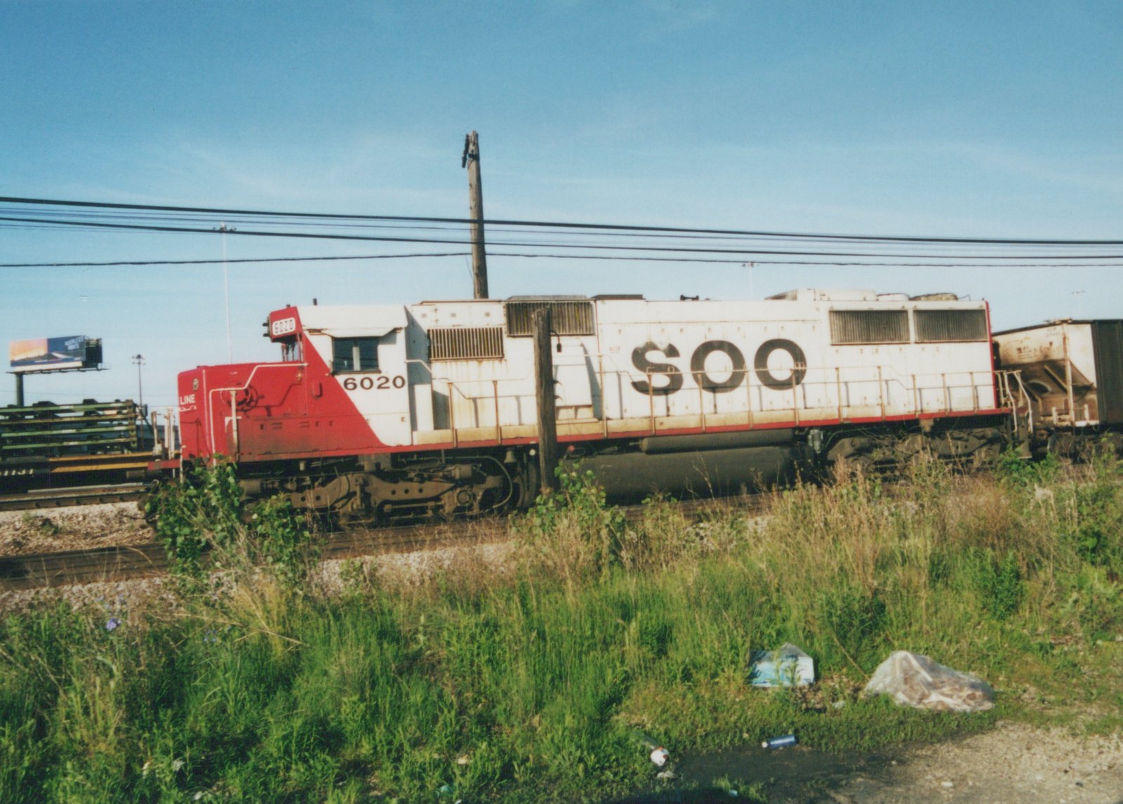 A red and white diesel locomotive with 'SOO' lettering on its side and the number 6020 on the cab and front, sitting on tracks in a grassy yard under a clear blue sky.