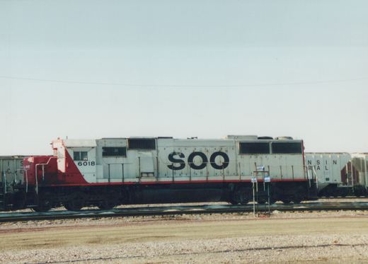 A side view of a white and red SOO Line diesel locomotive numbered 6018, parked on tracks with freight cars nearby under an overcast sky.