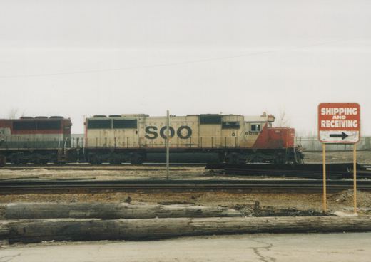 Two SOO Line diesel locomotives, numbers 6018 and 8301, are parked on parallel tracks in a rail yard under an overcast sky, with a 'SHIPPING AND RECEIVING' sign in the foreground.