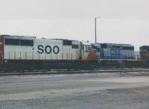 A side view of a white SOO locomotive numbered 6017 parked in a gravel yard next to a blue CSX locomotive and another SOO unit, all under a gray sky.